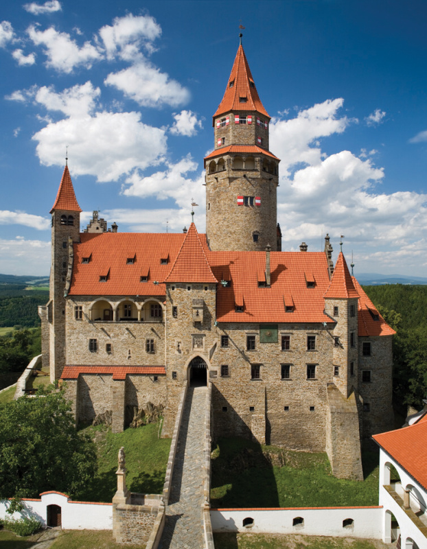 Středověký hrad s červenými střechami a věží na pozadí modré oblohy s bílými mraky.